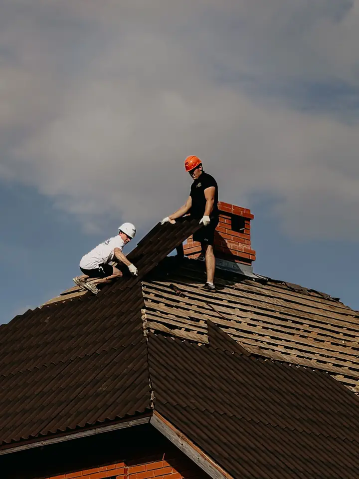 Two roofers in hard hats stripping old tile from a steep gable roof beside a brick chimney during a roof replacement in Kent, WA